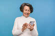 © InsideCreativeHouse - Happy aged caucasian woman grandmother holding smartphone using mobile online apps, browsing. Smiling old elderly lady texting sms message chatting with family on phone isolated over blue background.