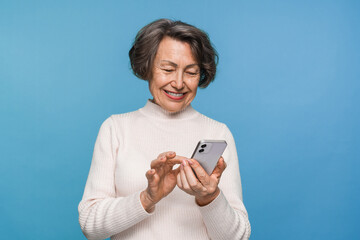 Happy aged caucasian woman grandmother holding smartphone using mobile online apps, browsing. Smiling old elderly lady texting sms message chatting with family on phone isolated over blue background.