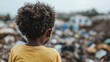 © svastix - A poignant image of a child with curly hair clad in a yellow shirt, seen from behind while observing a large pile of waste, symbolizing the harsh realities of poverty and wastefulness.