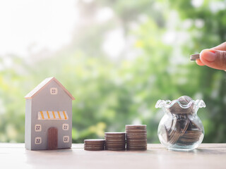 Poster - Close up hand putting coin into glass bottle, stack of coins and Miniature house.  The concept of saving money for house, Property investment, House mortgage, Real estate.