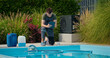© StockMediaSeller - Pool technician checking data on a tablet beside a swimming pool with chemical containers and lush greenery.