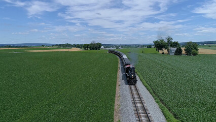 An Aerial view vintage steam locomotive moves along scenic tracks, surrounded by expansive green fields and towering trees, showcasing the charm of rural landscapes on a clear day.