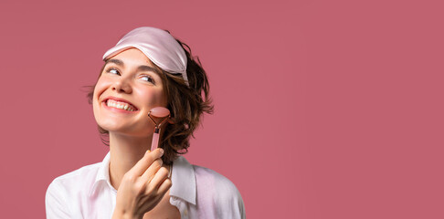 Young woman with rose quartz stone roller on pink background. Facial self care, morning beauty rituals, cosmetology, anti aging and anti-wrinkle treatment