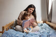 © Claudia - Mamá con bebé de un año sentado en sus piernas, ella le está leyendo un cuento, ellos están sentados sobre una cama. Actividad madre e hijo