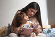 © Claudia - Mamá con bebé de un año sentado en sus piernas, ella le está leyendo un cuento, ellos están sentados sobre una cama. Actividad madre e hijo