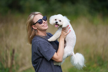 Portrait of smiling young woman kiss dog in Natural Park. Dog lover woman wearing blue dress with dog during good day. Concept of friendship and beautiful girl with a small dog