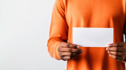 Person in an orange sweater holding a blank white card against a neutral background, emphasizing copy space and presentation concept.