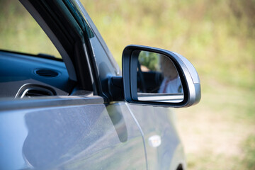 Close-up view of gray side mirror (or door mirror) of car in countryside in a sunny summer day. Blurred female driver reflects in mirror surface. Automobile industry theme.