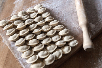 Raw dumplings were laid out on a wooden board for cooking. Next to it is a rolling pin for rolling out the dough. The filling is minced meat, potatoes, cheese or cottage cheese.