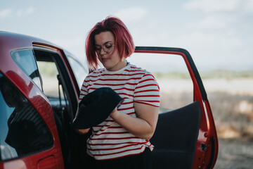 Woman with red hair and striped shirt getting ready for a road trip adventure. Preparing by her red car in a beautiful natural setting.