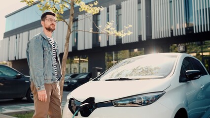 Handsome Caucasian young man standing outdoors in city putting charger cable into his electric car. EV owner charging his auto at charger station on street. Elecrtocar. Electric vehicle. Slow motion