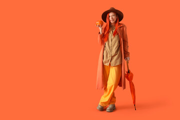 Poster - Beautiful young happy woman in autumn outfit with umbrella and pumpkin on orange background