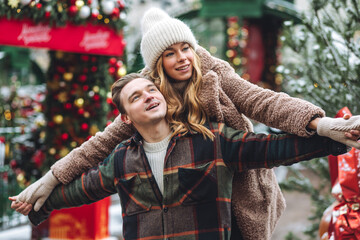 Concept of celebrating Christmas outdoors. Beautiful young loving couple enjoying winter holiday on decorated holiday market. Walking, kissing, hugging. String bag with tangerine, fir tree branches