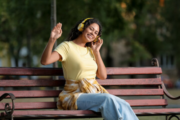 Wall Mural - Beautiful young African-American woman in headphones sitting on bench in park