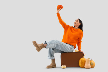 Beautiful young happy woman with suitcase and many pumpkins sitting on grey background