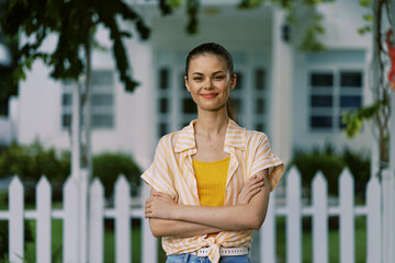 Young woman standing confidently in front of a white picket fence, wearing a striped shirt and a yellow top, with a bright smile and a friendly demeanor