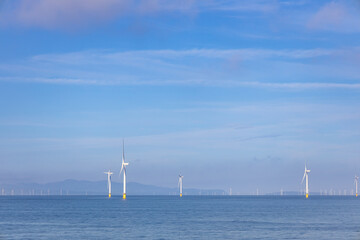 Abstract ethereal view of beautiful giant wind turbines at sea with patchy fog blue sky