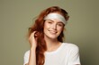 © Henry - Young woman with green eye patches on her face, isolated against a beige background. Close-up portrait of a ginger-haired girl wearing a T-shirt and looking away while smiling, applying a cosmetic gel