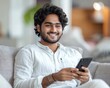 © Nattapat - Joyful young Indian man sitting on sofa, looking at mobile phone screen with a happy expression, celebrating success, hand in yes pose, comfortable home atmosphere