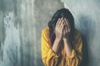 © Svetlana - A young girl in a yellow shirt sits against a wall, hiding her face in despair while struggling with fear and overwhelming emotions
