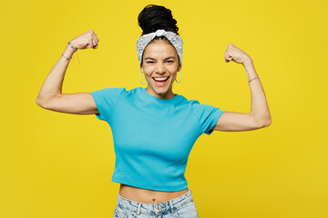 Wall Mural - Young Latin woman she wears blue top t-shirt headband casual clothes showing biceps muscles on hand demonstrating strength power isolated on plain yellow background studio portrait. Lifestyle concept.