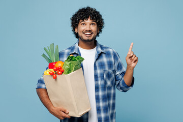 Wall Mural - Young happy Indian man wear shirt hold brown craft bag for takeaway mock up with food products point finger overhead on area isolated on plain blue background Delivery service from shop or restaurant