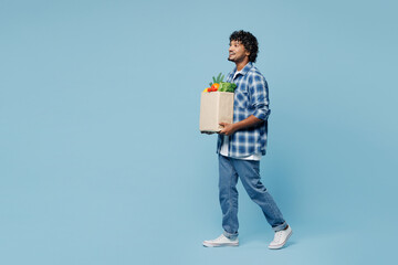 Wall Mural - Full body side profile view fun young Indian man wear shirt hold craft bag for takeaway mock up with food products walk go isolated on plain blue background. Delivery service from shop or restaurant.