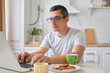 © sementsova321 - Focused young man wearing glasses using laptop typing on keyboard writing email or message chatting shopping successful freelancer working online on computer sitting in modern kitchen