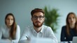 © kokoson6 - A young man with a beard and glasses looks intently at the camera during a business meeting or lecture. Two female colleagues are in the background.