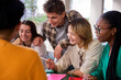 © CarlosBarquero - Group of international young university students meeting together looking homework in campus study. Diverse Gen z high school people smiling pointing to syllabus taking notes sitting at faculty table