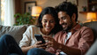 © mischenko - indian couple smiling and looking at phone sitting on sofa at home