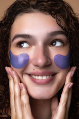 A close-up beauty portrait of a smiling young girl with curly hair, wearing purple under eye patches. She gently touches her face showing a skin care routine .