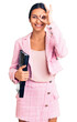 © Krakenimages.com - Young beautiful latin girl wearing business clothes holding binder smiling happy doing ok sign with hand on eye looking through fingers