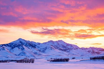 Canvas Print - A fiery sunset paints the sky above snow-capped mountains, casting a warm glow on the winter landscape.