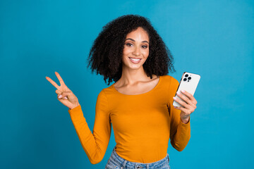 Poster - Photo portrait of attractive young woman hold device show v-sign dressed stylish yellow clothes isolated on blue color background