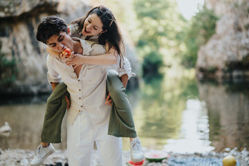 Poster - A cheerful young couple having fun by the river, with the woman playfully on the man's back, enjoying a sunny outdoor day with laughter and fresh fruit.