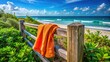 © Achiraya - Vibrant orange beach towel with a subtle texture, gently rumpled and draped over a weathered wooden beach fence, surrounded by lush greenery and ocean views.