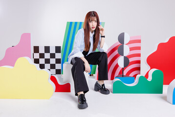 Wall Mural - Fashion photo young confident brunette woman model Asian beautiful female wearing striped blue shirt and black pants posing looking at camera, sitting on white box posing in studio colorful background