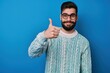 © Vadym - Young handsome bearded man in casual sweater, glasses stands on blue background. Gives happy thumbs up gesture with right hand. Man looks at camera with approving expression, showing success,