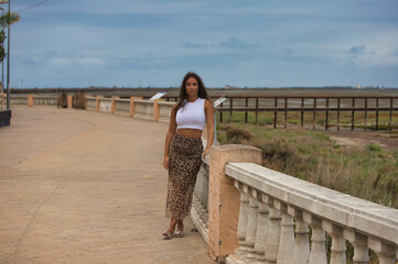 Wall Mural - Latin woman, young and beautiful dressed in a white shirt and tiger print skirt leaning on a stone railing. In the background the salt mine and the city of Cadiz on the horizon.