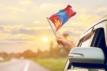 Waving the Mongolia flag against the sunrise or sunset from a car driving along a country road. Holding the Mongolia flag, traveling by car, on a weekend trip.