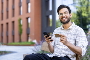 Wall Mural - Portrait of a young Indian man holding a credit card and a mobile phone, sitting on a bench outside a building and looking at the camera