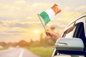 Waving the Ireland flag against the sunrise or sunset from a car driving along a country road. Holding the Ireland flag, traveling by car, on a weekend trip.