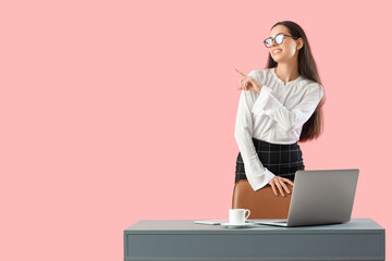 Wall Mural - Pretty young female programmer standing near table with laptop and pointing somewhere on pink background