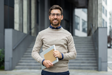 businessman standing outside office building holding laptop. smiling confidently, wearing glasses an