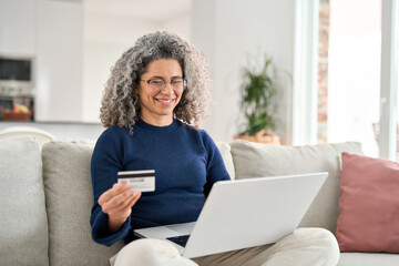 Mature lady customer looking at computer buying online doing ecommerce shopping on website making bank payments. Happy curly middle aged woman using laptop holding credit card sitting on couch at home