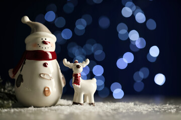 Wall Mural - Cute decorative snowman and deer on artificial snow against blurred lights. Space for text