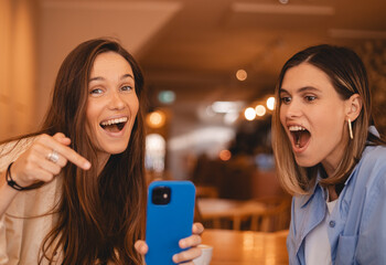 Portrait of excited impressed two  women sisters friends fellows use devices recommend select choose decide discount options, point finger on phone, screaming wow, open mouth, omg, sitting in cafe.