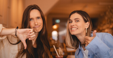 Two women sisters friends rest in cafe, she show like and dislike, thumb up and thumb down, disagreement and agreement expression. Crazy conflict. Different opinion. 