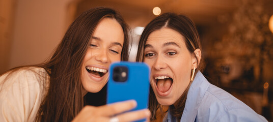 Overjoyed happy excited two women say wow, glad to receive text message informing salary, rejoices good news, stares at mobile phone, gestures actively from happiness, video call. Girls rest in cafe